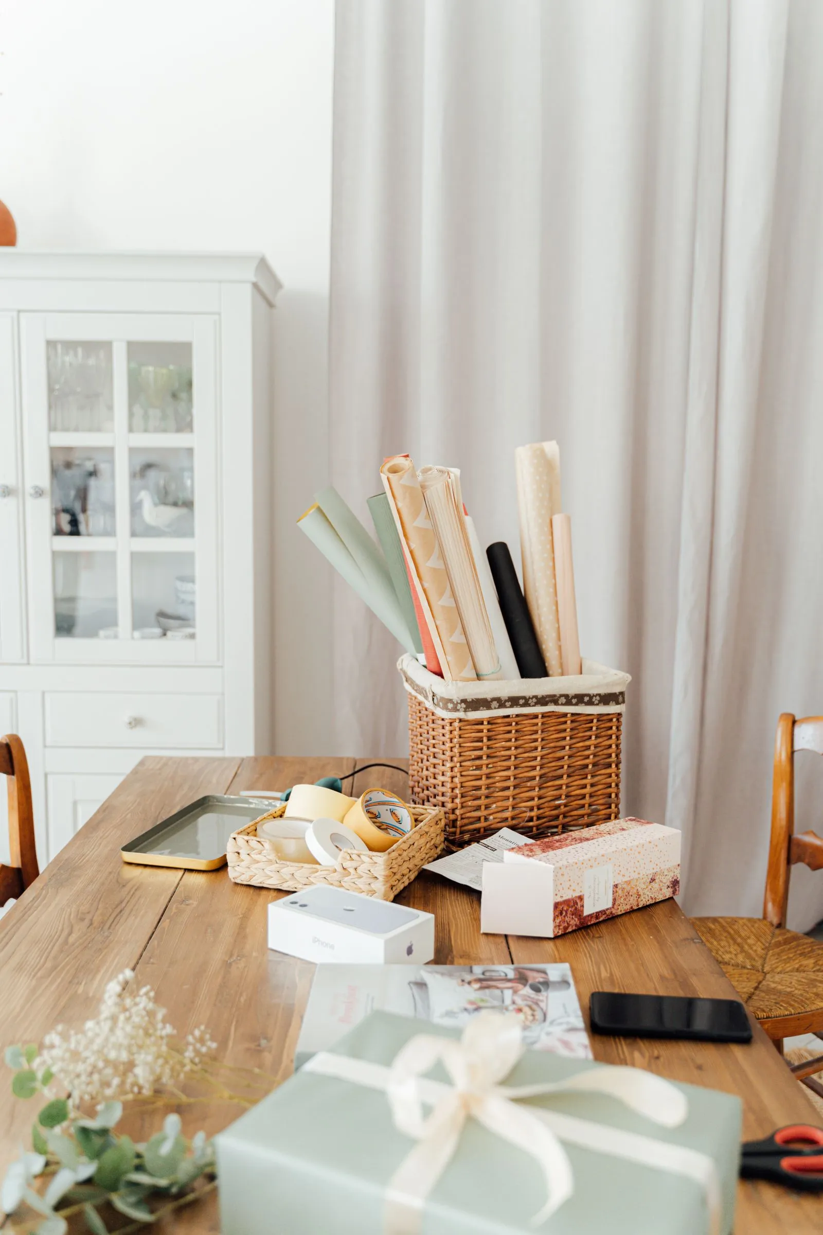 Rolls of wrapping paper standing upright in a fabric-lined wicker basket on a wooden dining table, with rolls of tape in a small woven tray beside it