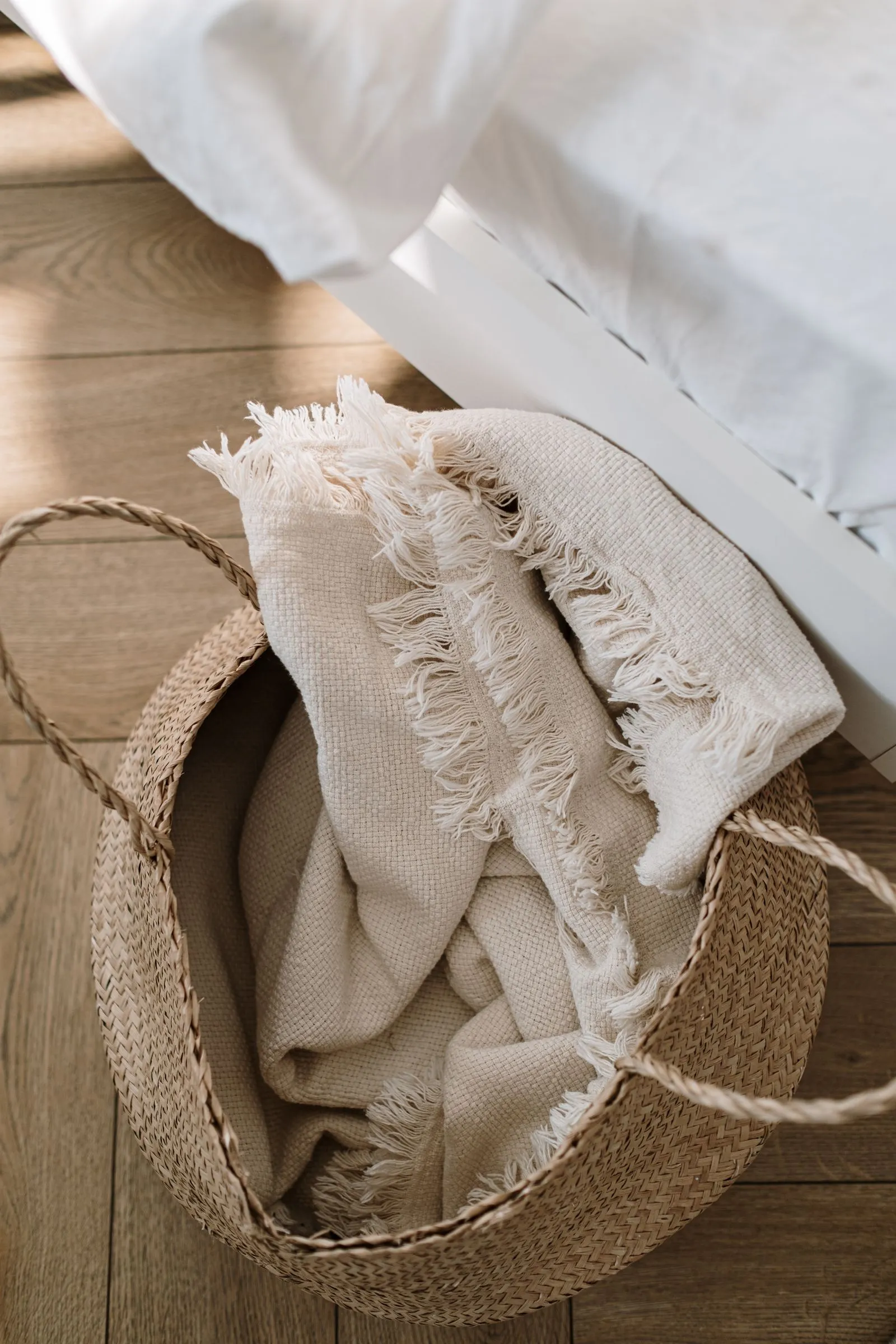 Woven storage basket holding a folded ivory throw blanket sitting on the floor beside a white bed frame
