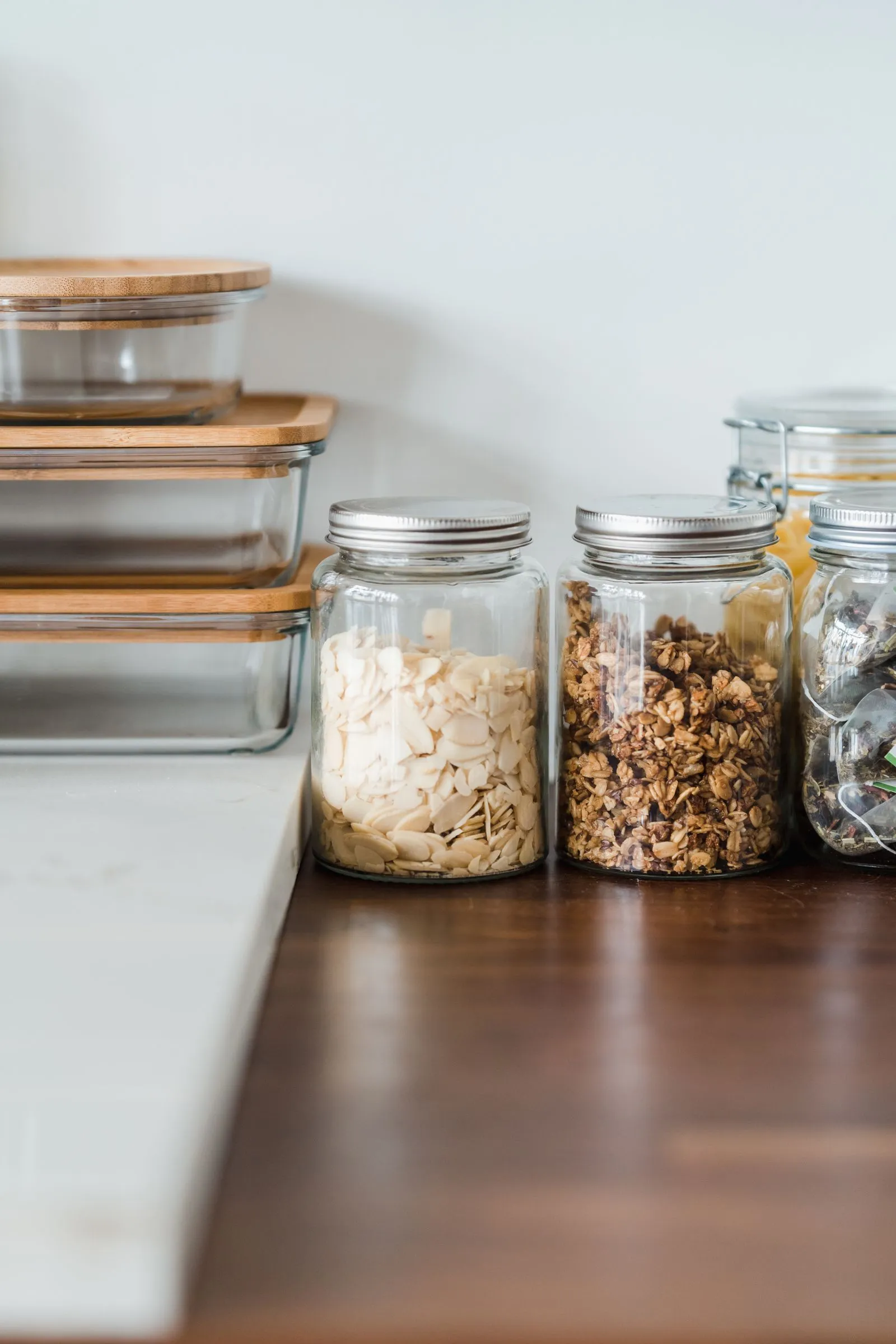 Glass jars filled with almond slices, granola, and loose-leaf tea bags lined up on a wood counter next to stacked glass storage containers
