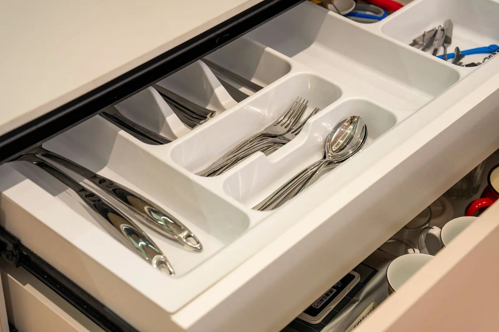 A white compartment tray inside an open kitchen drawer holding knives, forks, and spoons in separate slots, with a second divided tray of tools behind it