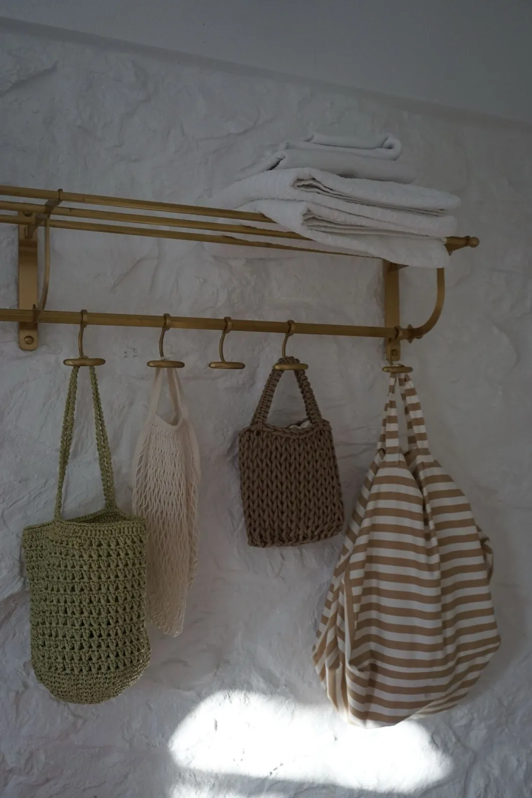 A row of gold wall hooks holding woven tote bags with folded towels stacked on the shelf above
