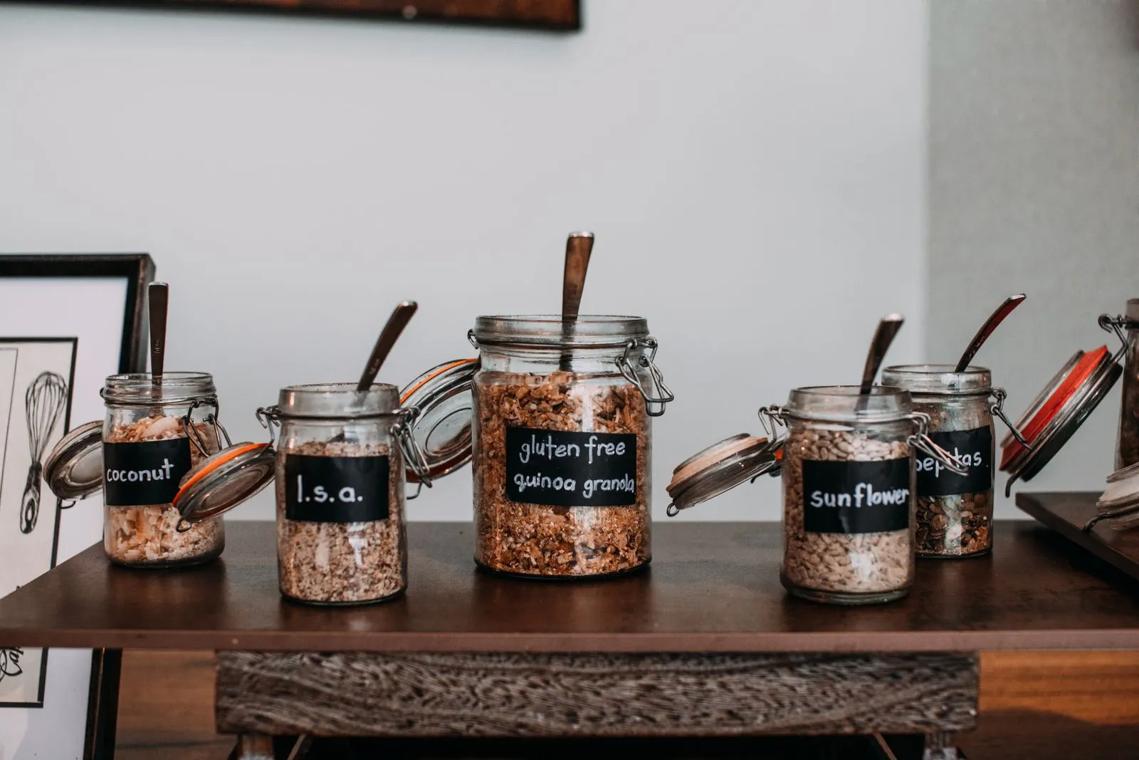 Glass jars with chalkboard-style labels reading coconut, l.s.a., gluten free quinoa granola, sunflower, and pepitas, lined up on a wooden table in a home kitchen