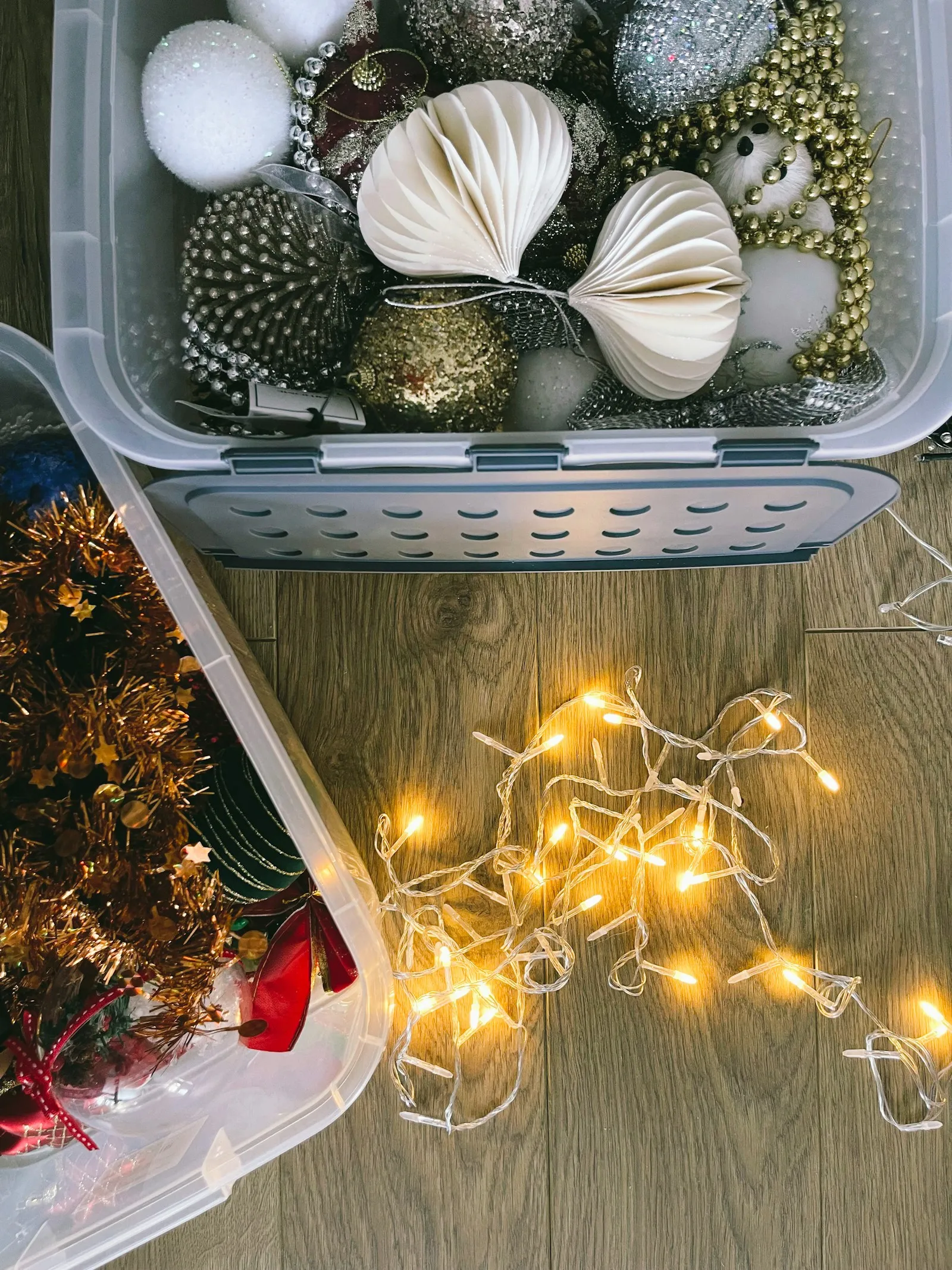 An open plastic storage bin of glittery ornaments and paper decorations on a wood floor next to a loose, tangled strand of lit string lights