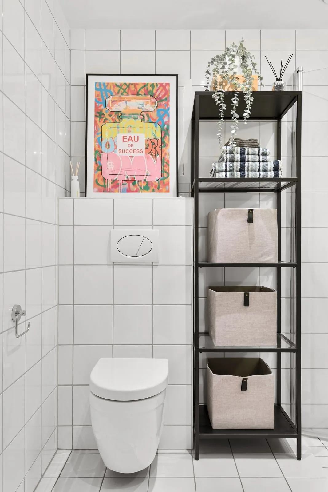 A narrow black metal shelving unit beside a wall-mounted toilet in a white-tiled bathroom, holding three fabric storage bins, folded towels, and a trailing plant on top