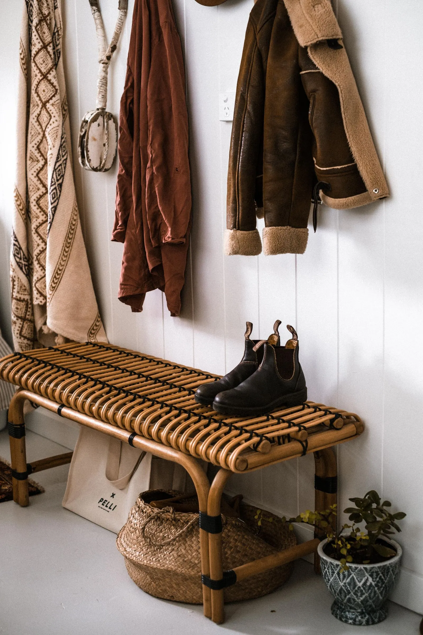Rattan bench with a pair of brown boots on the seat and a woven basket on an open shelf underneath, with coats hanging on the wall above