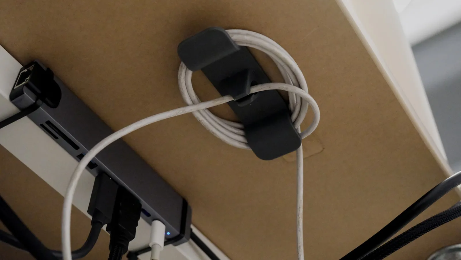A black adhesive cable clip mounted under a desk holding a coiled white cord next to a USB hub