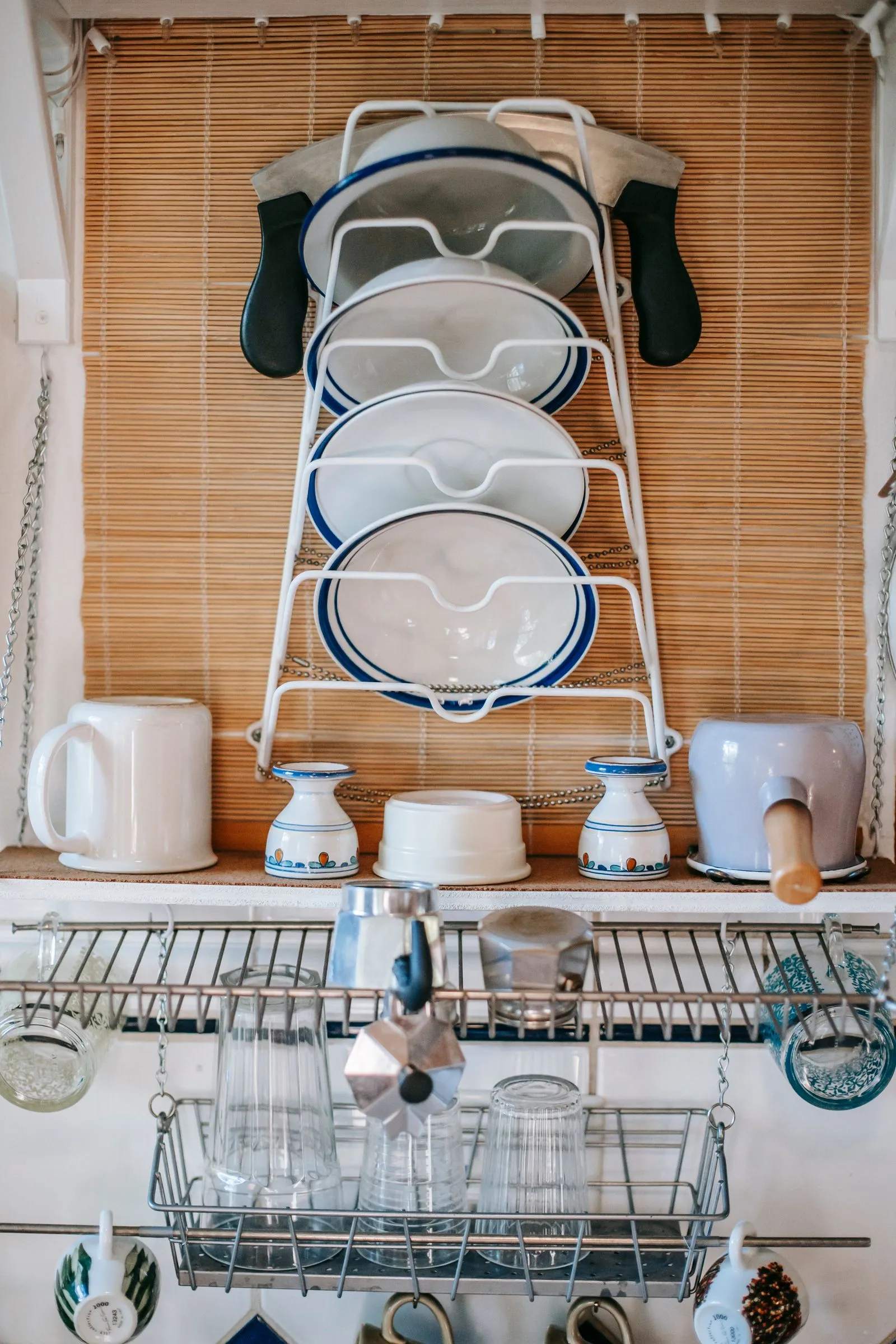 Wall-mounted metal lid rack holding pot lids above a shelf of mugs, jars, and dishware, with a wire basket rack below holding drinking glasses