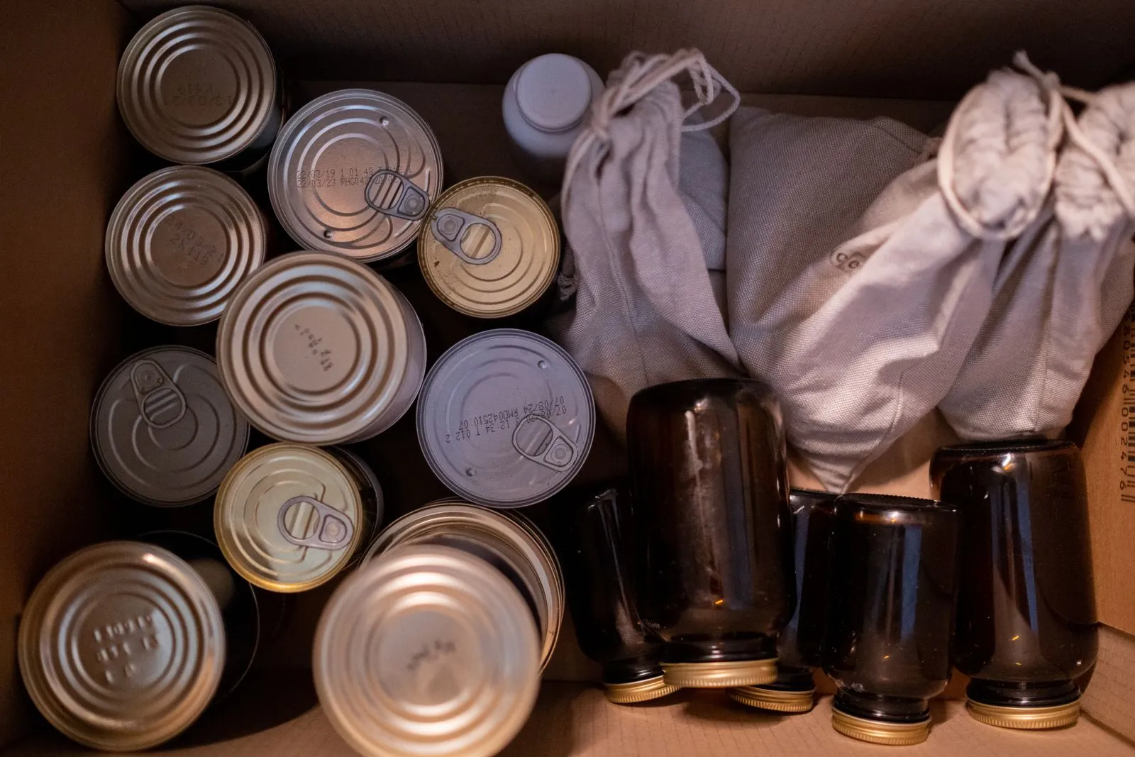 Overhead view of unlabeled canned goods and dark glass jars grouped inside a cardboard box alongside two tied cloth drawstring bags