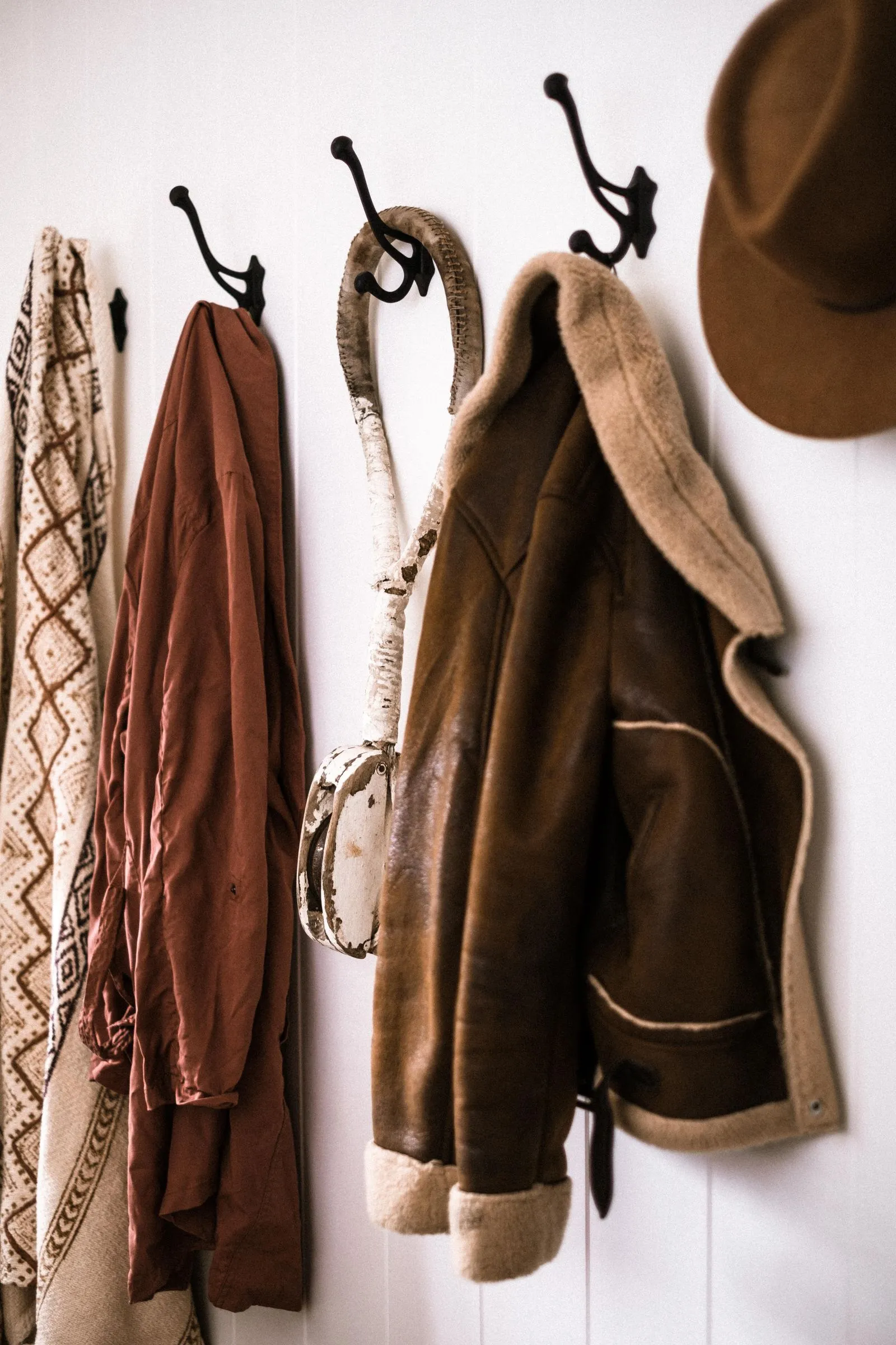 A row of four wall hooks holding a knit sweater, a rust-colored shirt, a shearling-lined leather jacket, and a felt hat