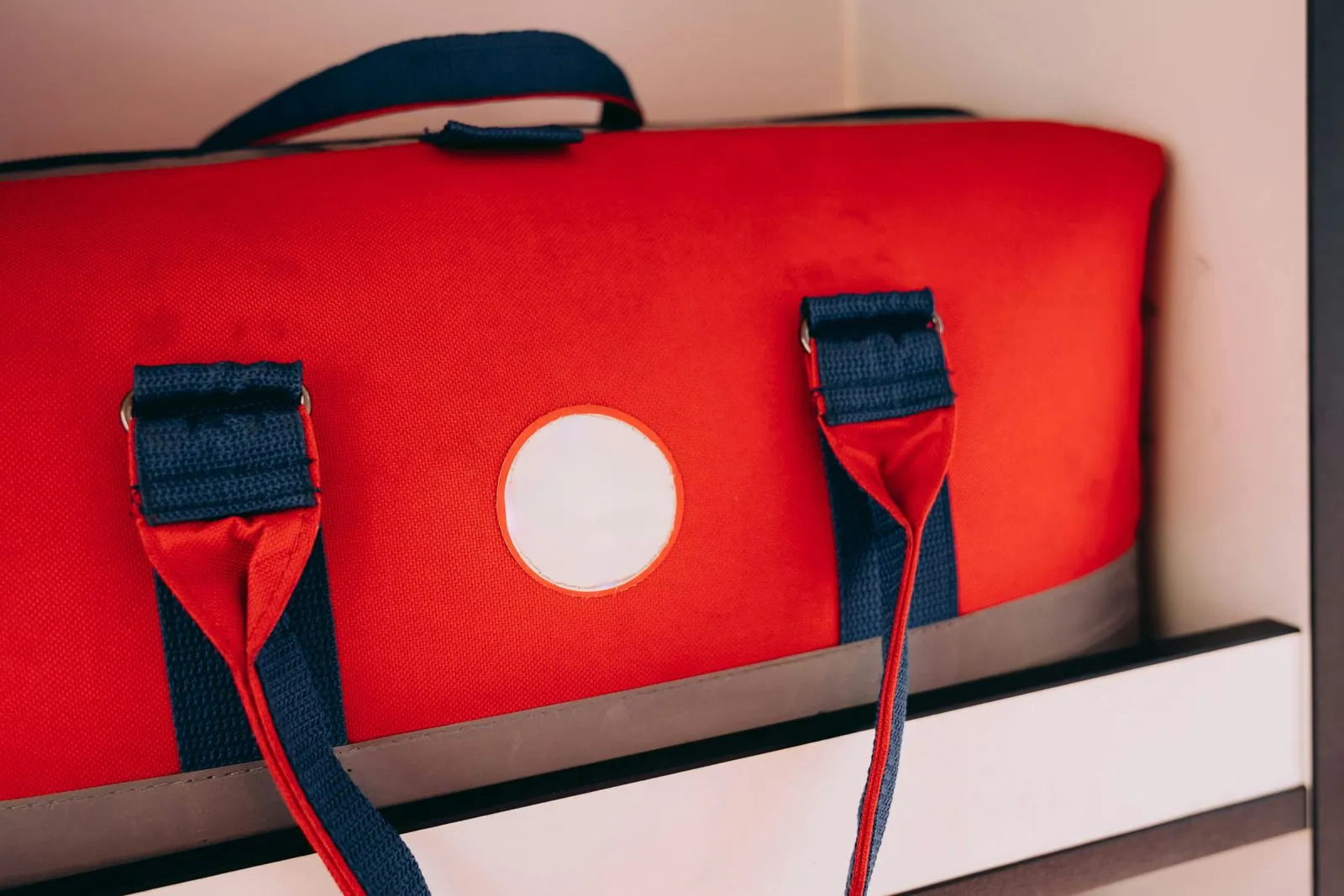 A red canvas duffel-style storage bag with navy straps sitting on a closet shelf