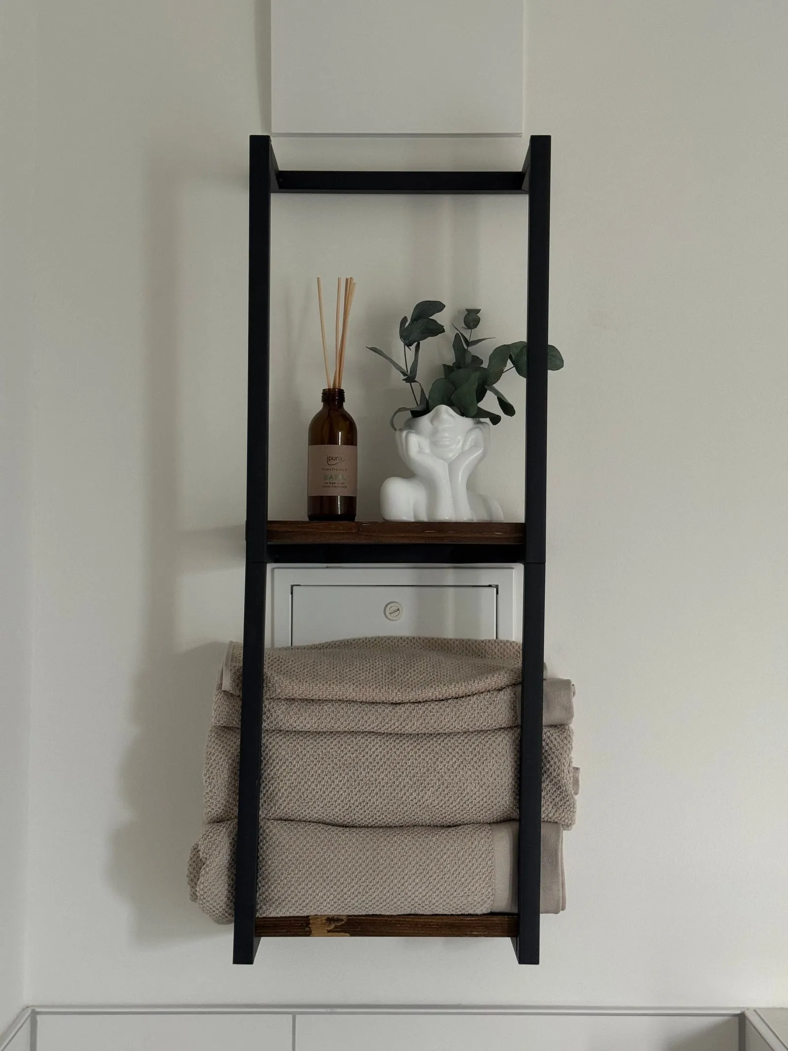 A black ladder shelf mounted on a bathroom wall, holding a reed diffuser and small vase on the top shelf and a stack of folded taupe towels on the bar below
