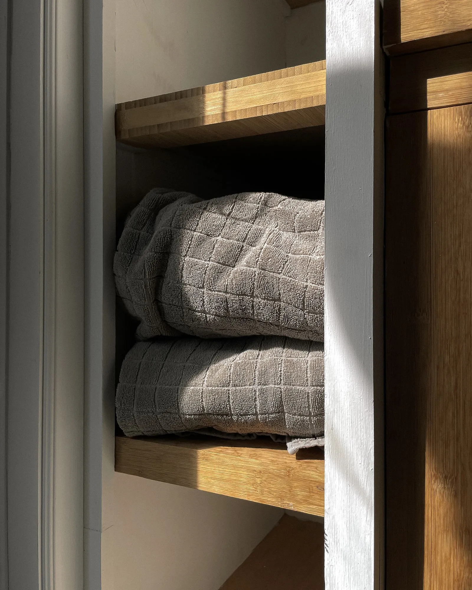 Two folded gray towels stacked squarely on a wooden shelf inside a sunlit closet nook