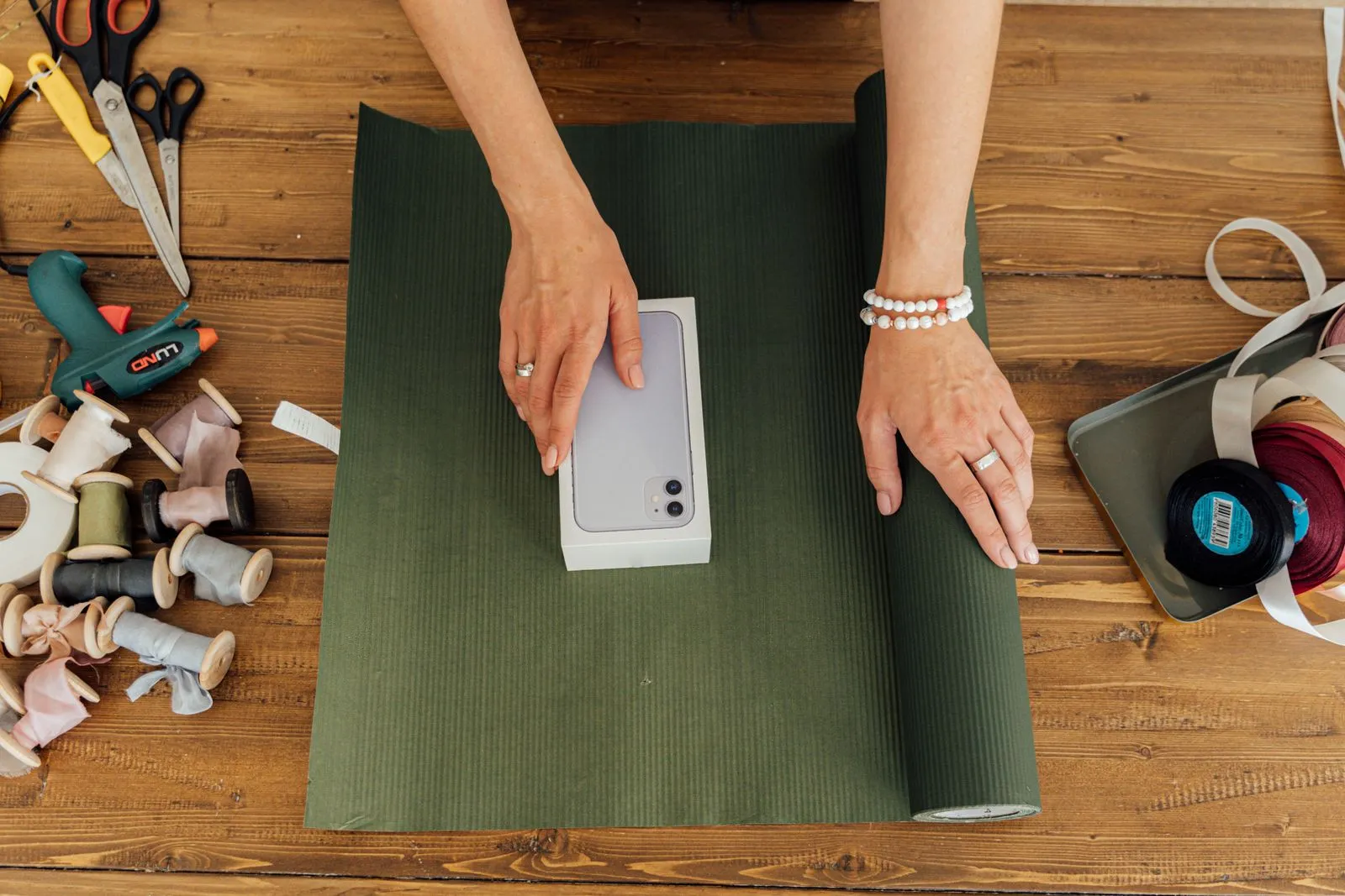 Hands positioning a small box on a sheet of green wrapping paper, with spools of ribbon, scissors, and a glue gun arranged on a wooden table nearby