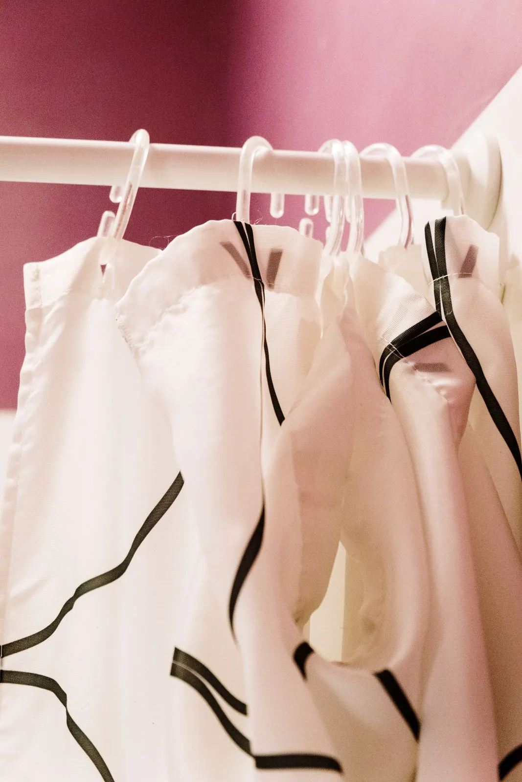 A white shower curtain with a black line pattern hanging from clear rings on a white rod against a pink wall