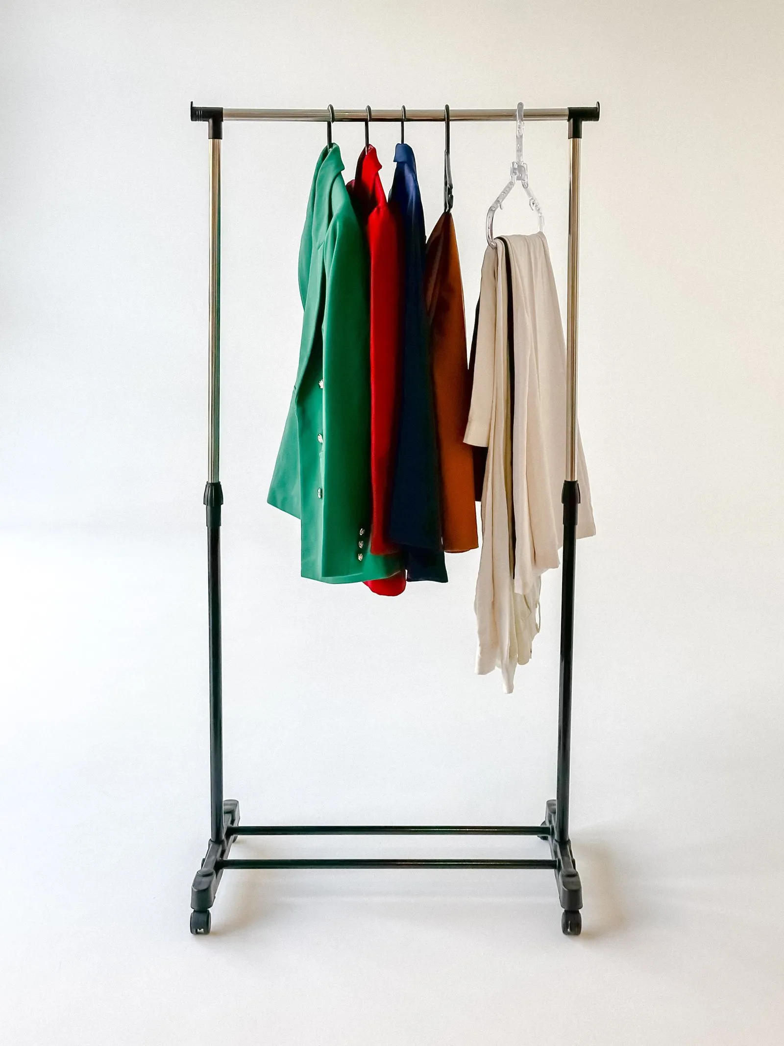 Empty wheeled clothing rack with a green blazer, red dress, navy jacket, rust-colored coat, and cream garment hanging from it against a plain background