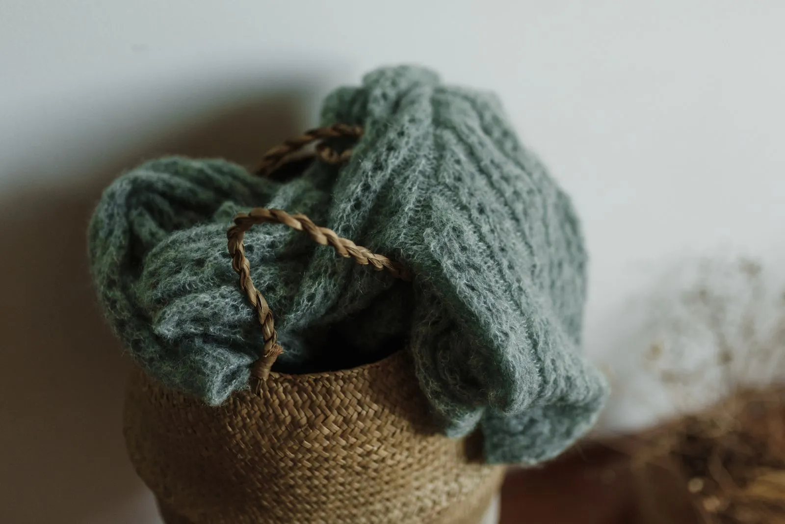A chunky sage-green cable-knit sweater folded over the rim of a woven seagrass basket with braided handles