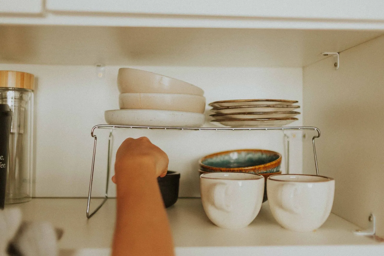 Wire shelf riser holding stacked bowls and plates above a row of mugs and small bowls inside a kitchen cabinet