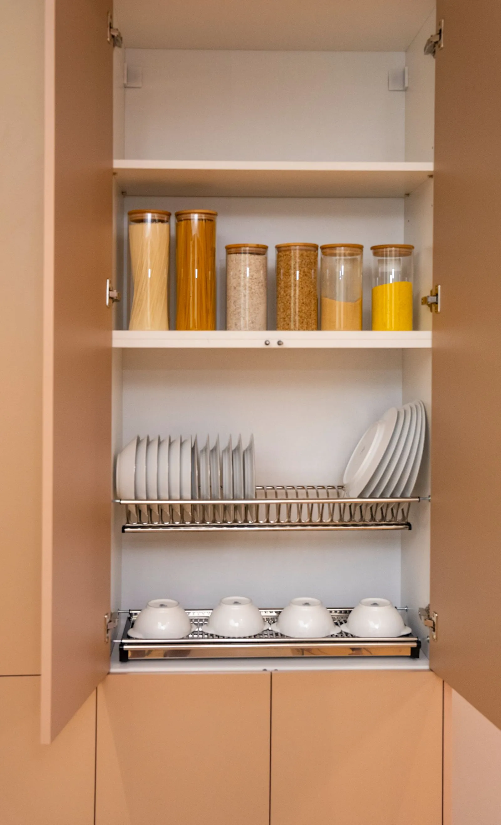 Open kitchen cabinet with glass canisters of pasta and grains on the top shelf, and two chrome pull-out wire racks below holding stacked plates and four cups with saucers