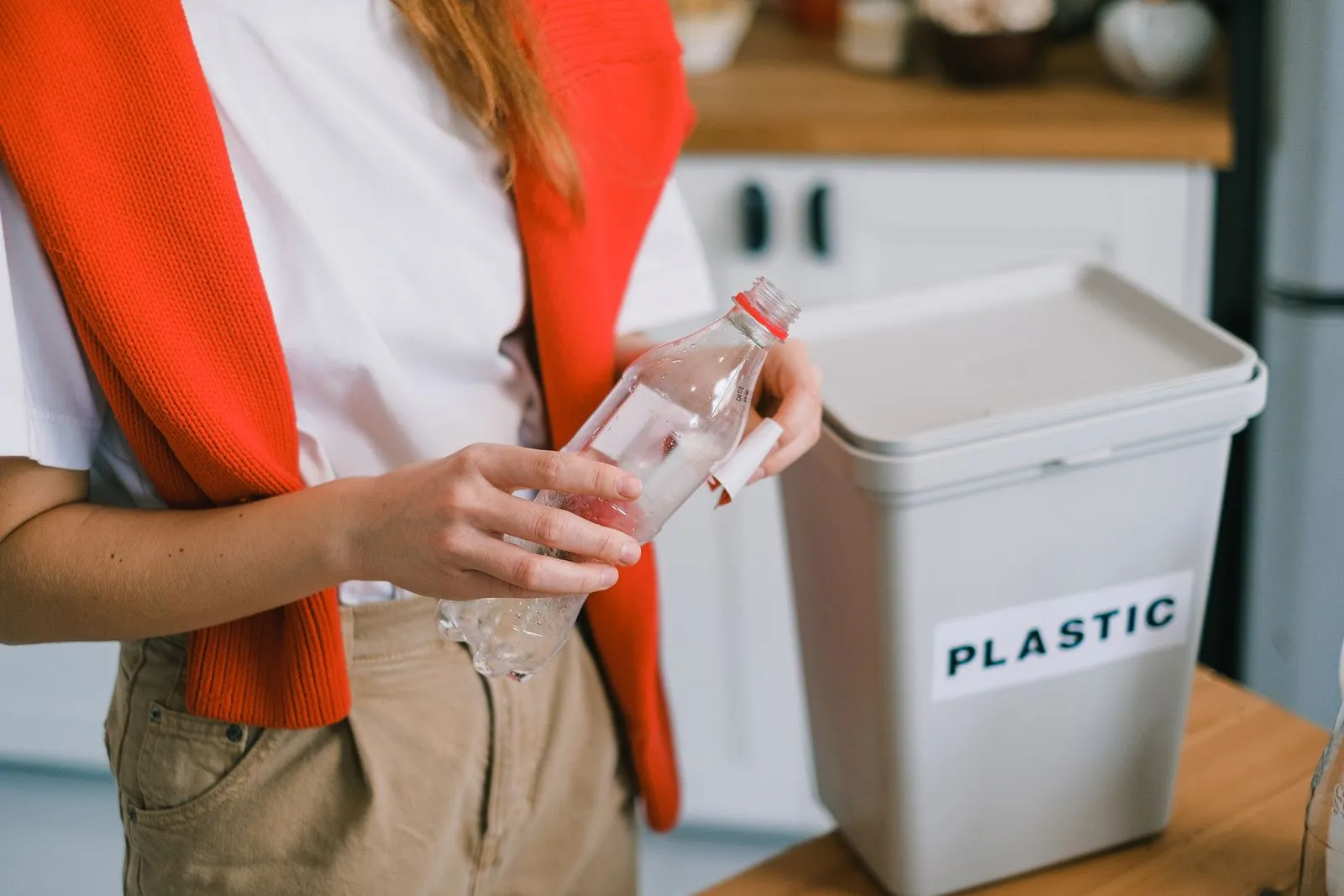 Person holding a plastic bottle next to a slim bin labeled PLASTIC on a kitchen counter