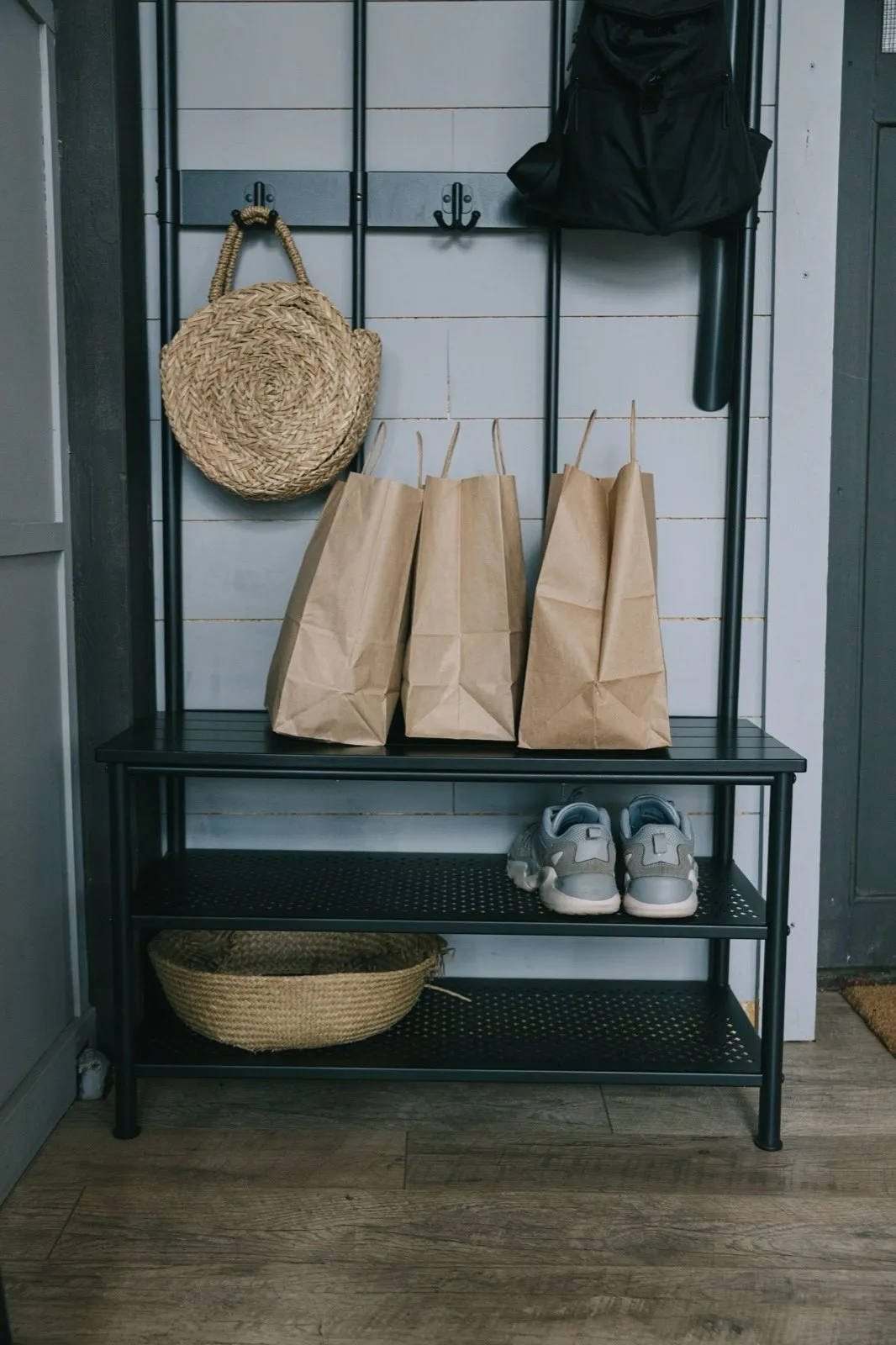 An entryway rack with a round woven bag and a backpack on wall hooks, paper shopping bags set on the bench top, and sneakers and a basket on the shelves below
