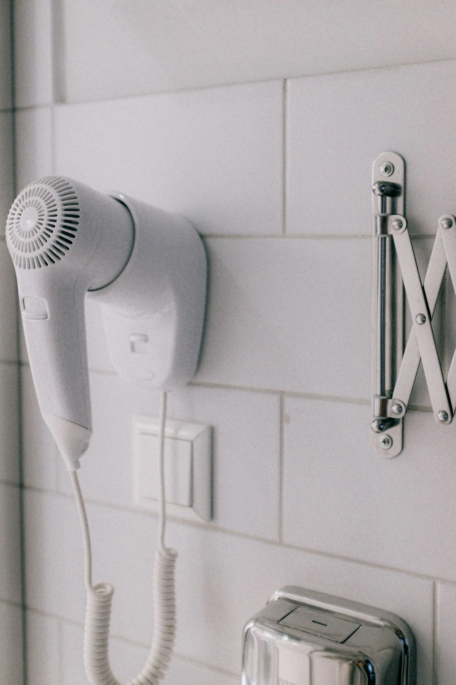 A wall-mounted hair dryer with its coiled cord hanging against white bathroom tile