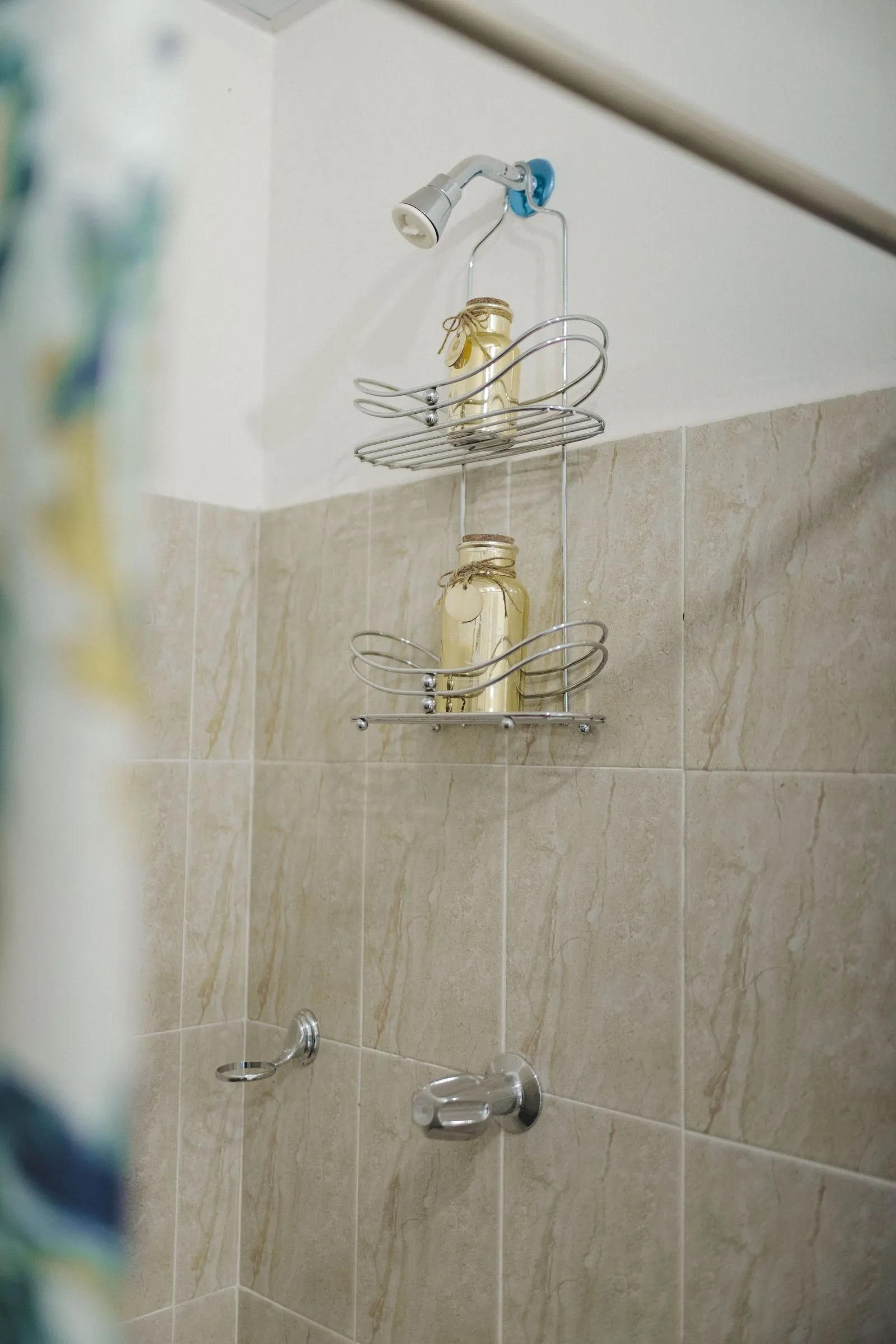 A two-tier chrome wire caddy hangs from hooks looped over a showerhead pipe, holding two corked glass bottles, against beige tiled shower walls