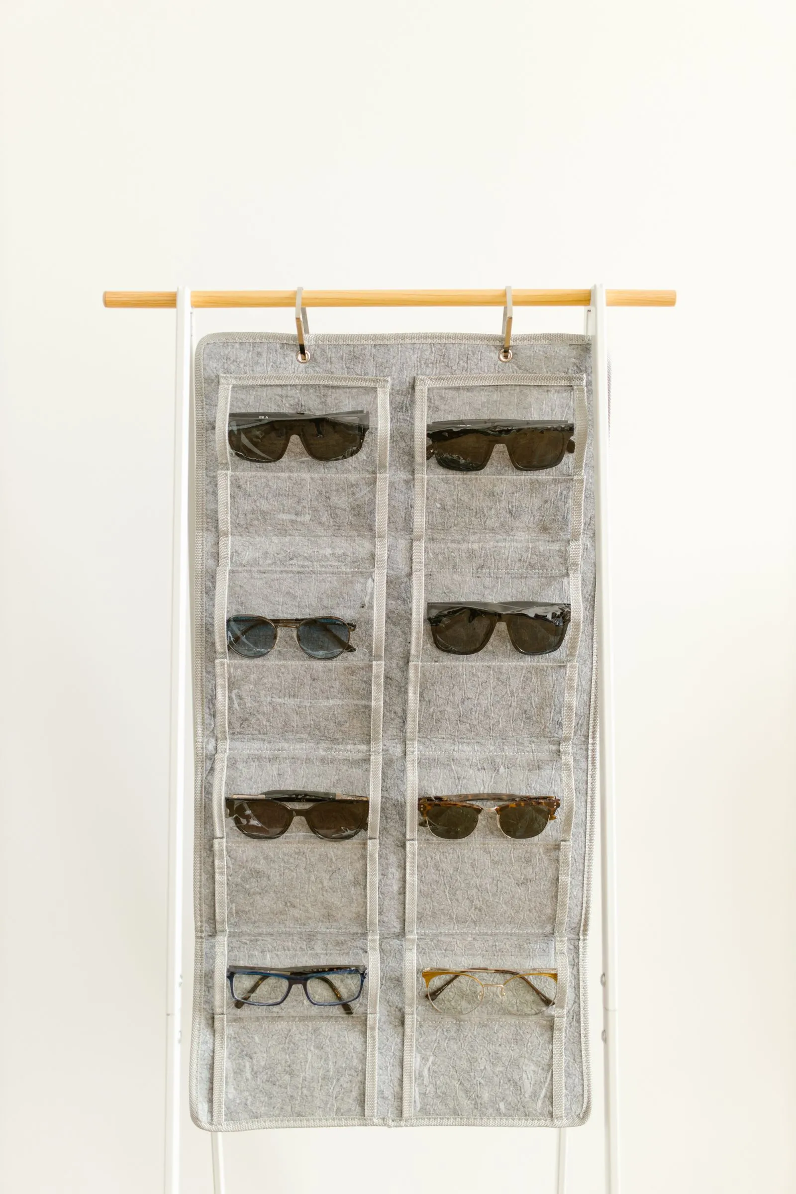 A gray felt pocket organizer with two rows of clear-front pockets hangs by loop straps over a wooden rod, each pocket holding a pair of sunglasses or eyeglasses