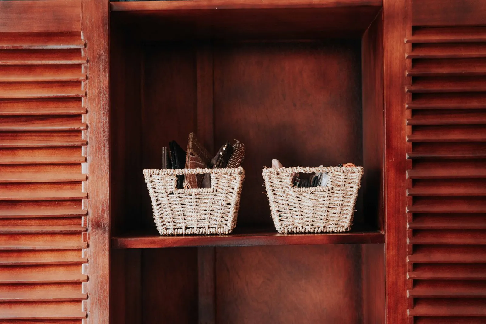 Two woven storage baskets sitting on a wooden shelf inside an open cabinet with slatted doors