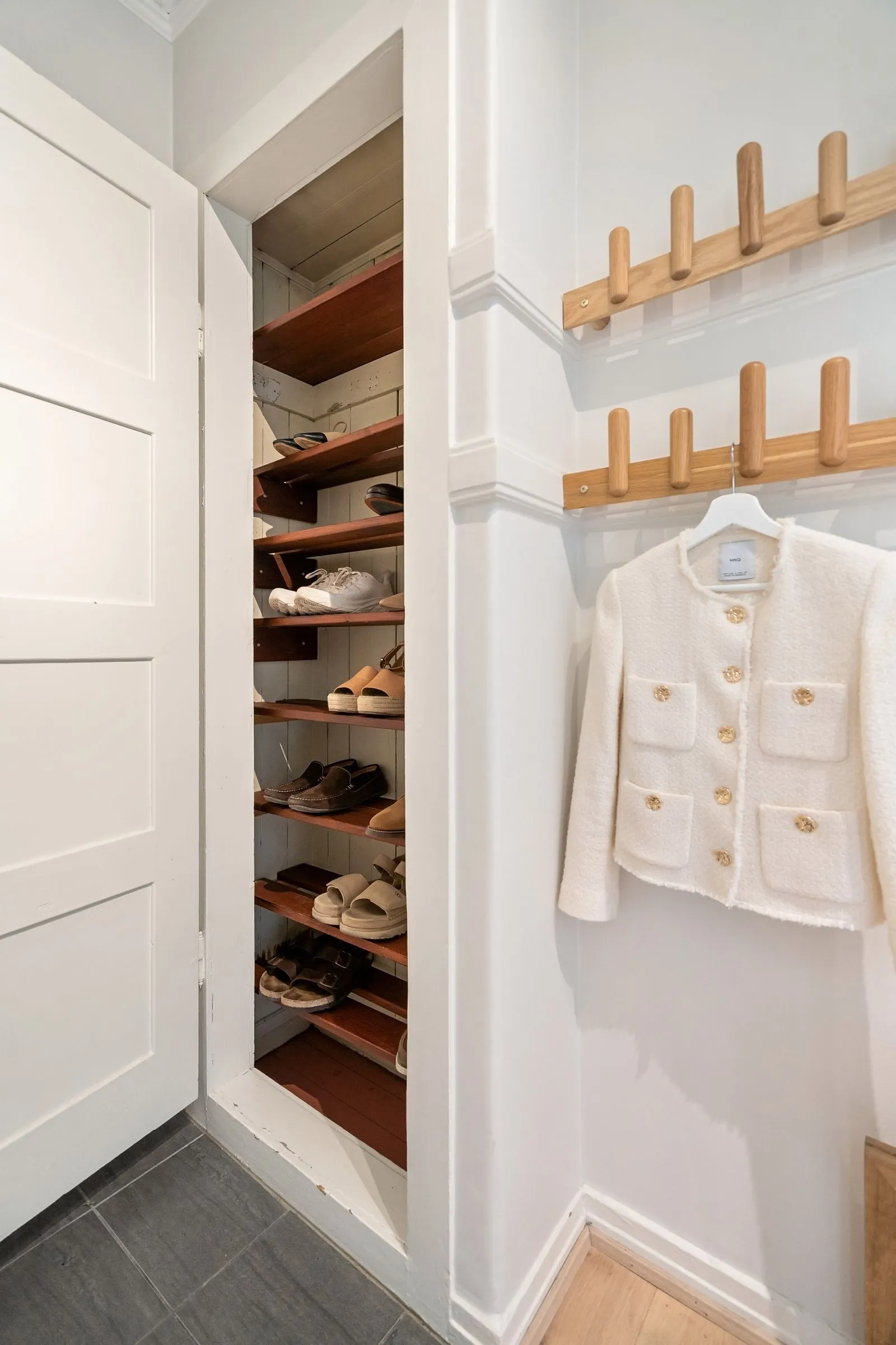A narrow closet with angled wooden shoe shelves holding sandals, sneakers, and loafers, with wooden peg rails and a white jacket on the wall beside it