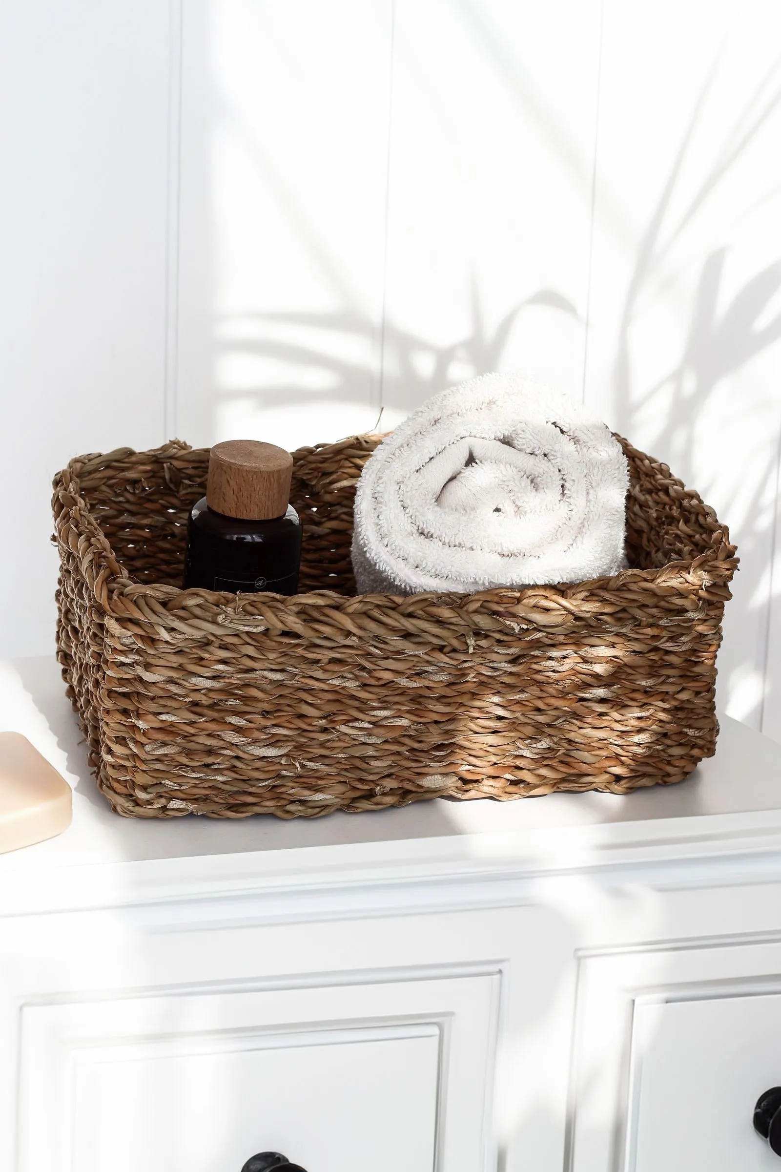 A woven basket holding a rolled towel and a bottle on a bright, sunlit cabinet