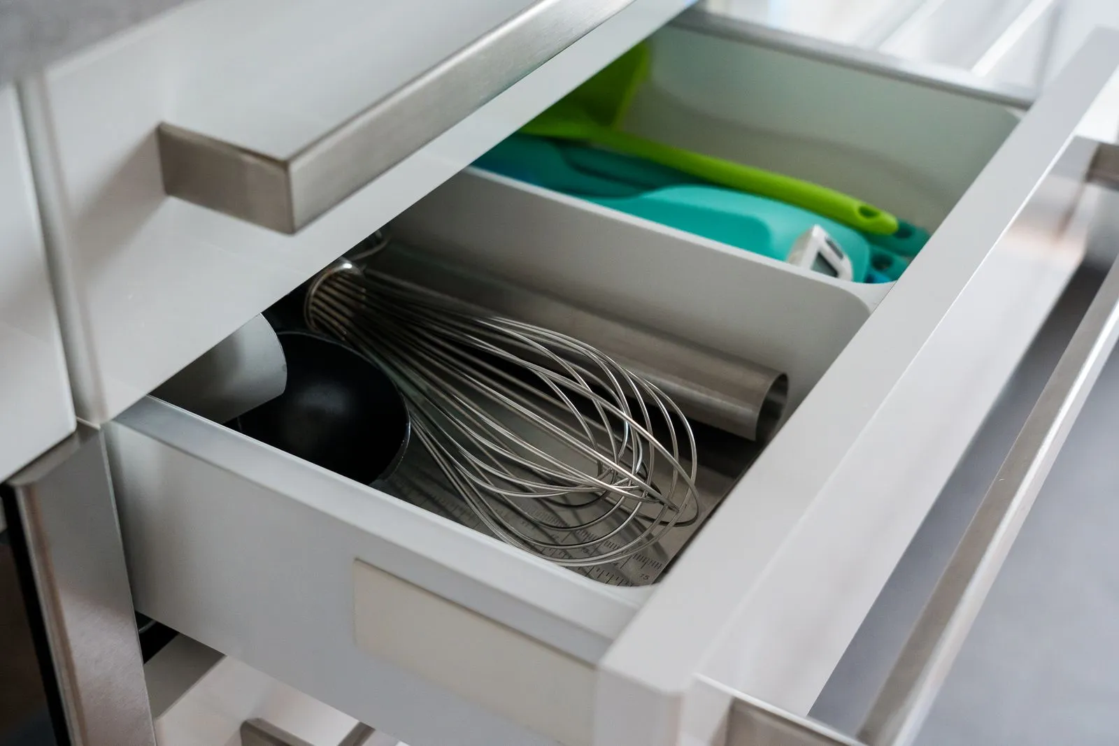 An open kitchen drawer split by a low divider, with a whisk and other metal tools in the front section and green silicone spatulas in the section behind