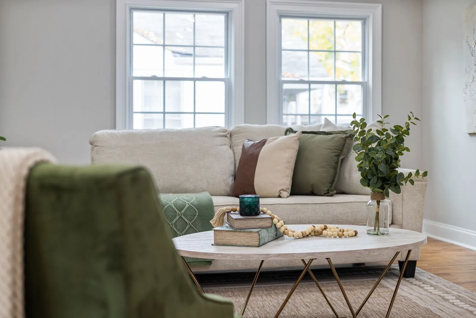 Oval light wood coffee table holding books, a candle, and a vase of eucalyptus in front of a cream sofa in a bright living room