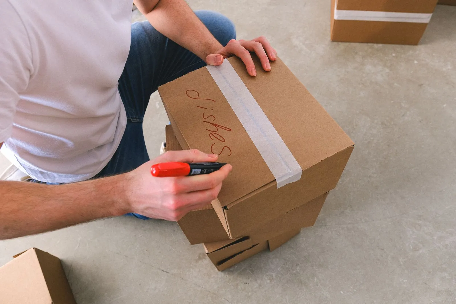 A hand writing a label in red marker on a taped cardboard storage box, with more boxes stacked nearby
