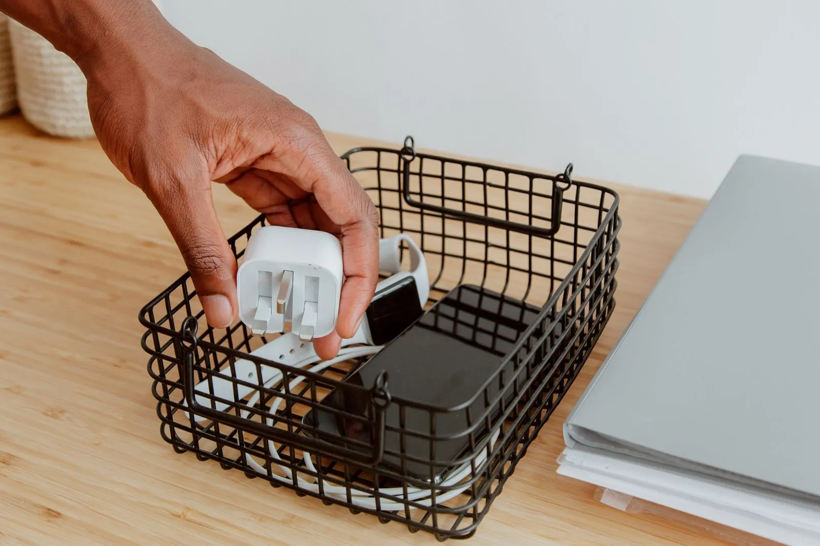 A hand placing a white charger brick into a small wire basket that already holds a coiled charging cable, a phone, and a watch