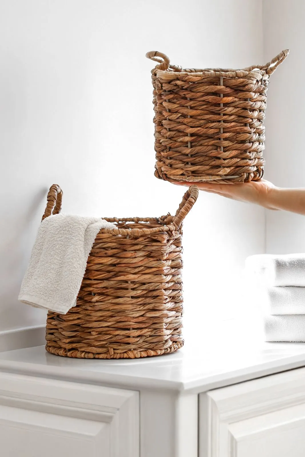 Two woven storage baskets on a white bathroom vanity counter, one lifted by hand, with a white hand towel draped over the lower basket and folded towels stacked alongside