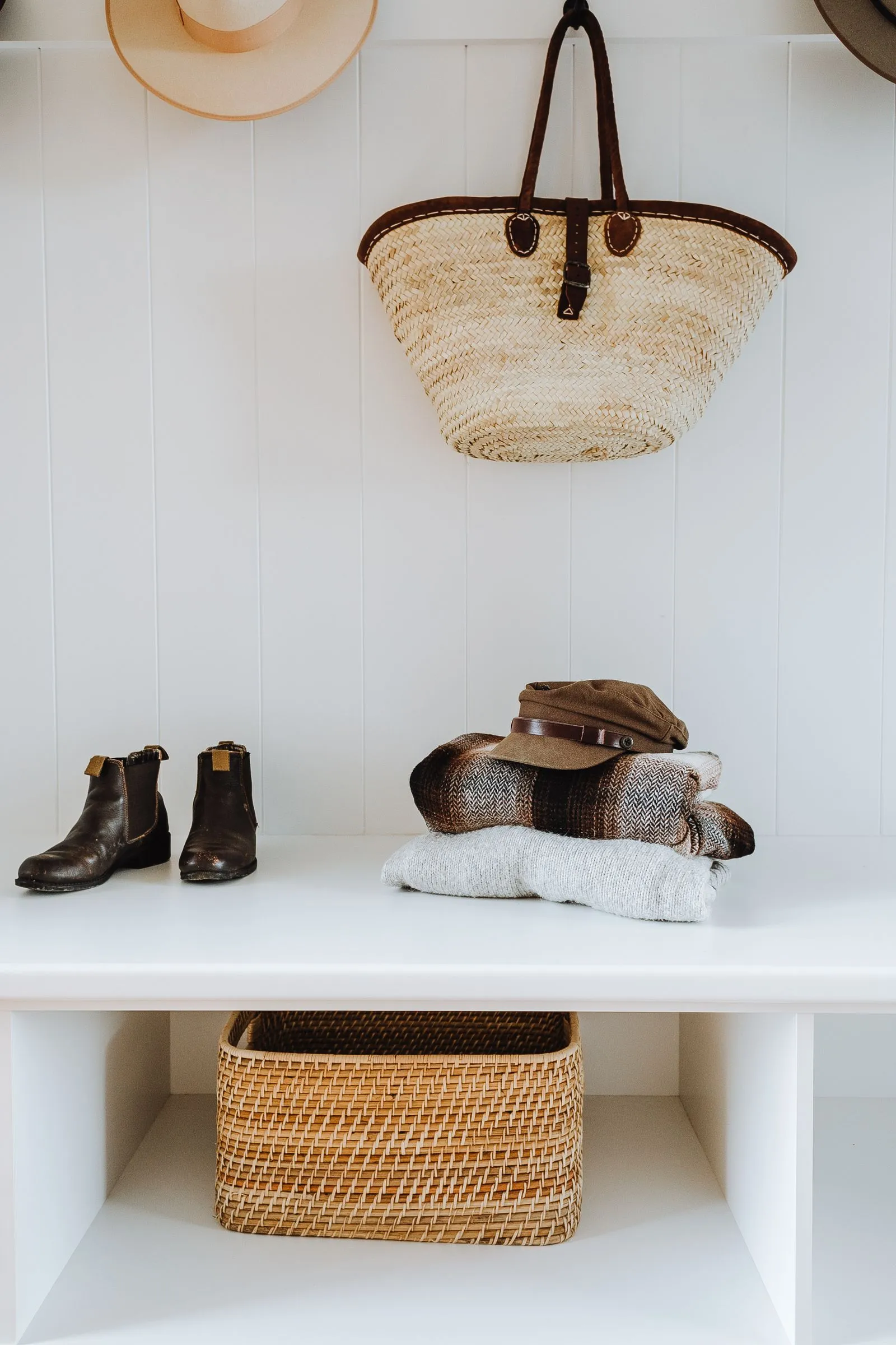 White entryway bench with brown boots and folded sweaters on top, a woven basket sitting in an open cubby underneath, and a hat and tote bag hanging on hooks above