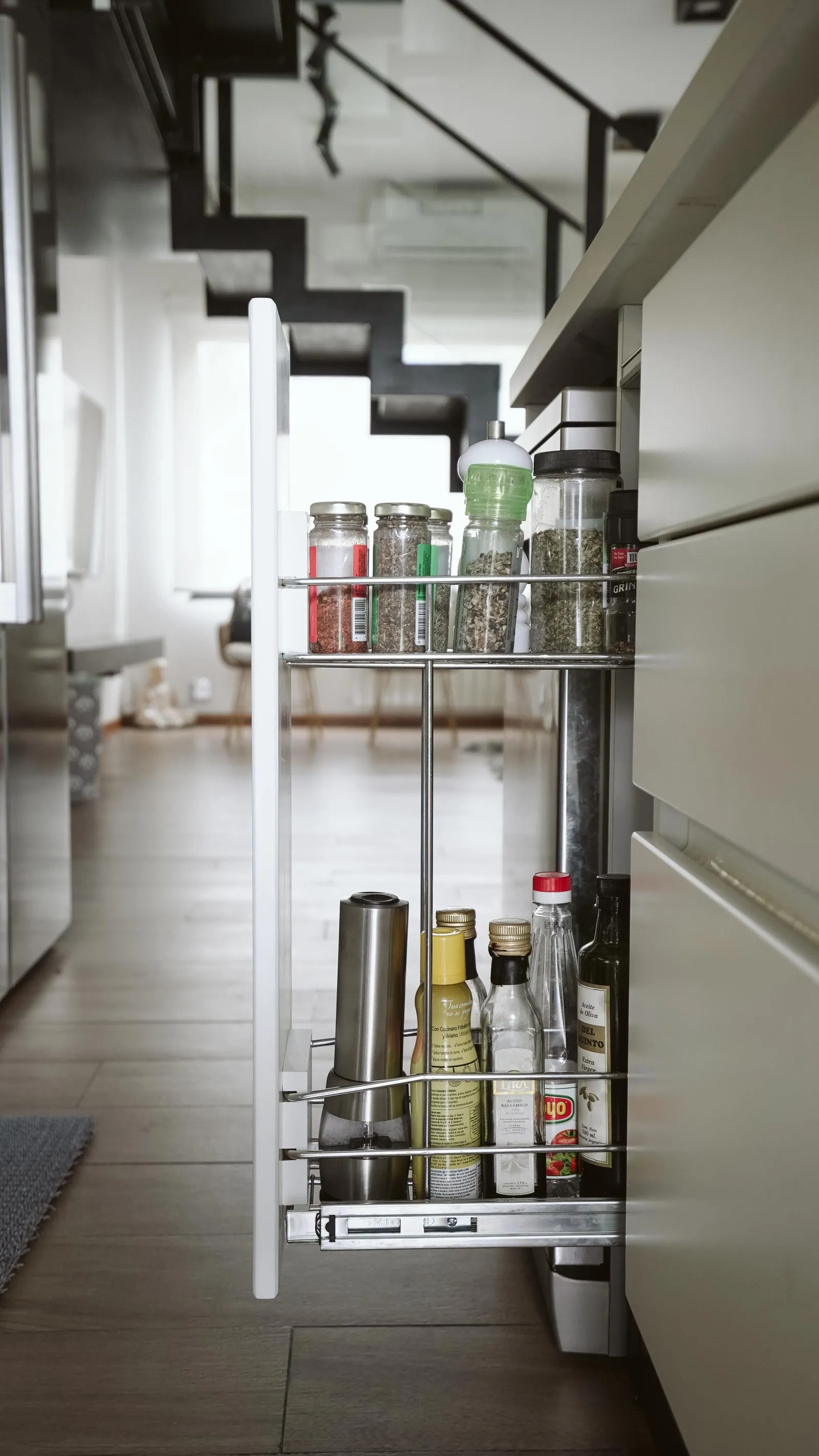 A narrow pull-out spice rack mounted to the inside of a slim cabinet, with small spice jars on the top tier and oil and vinegar bottles on the lower tier
