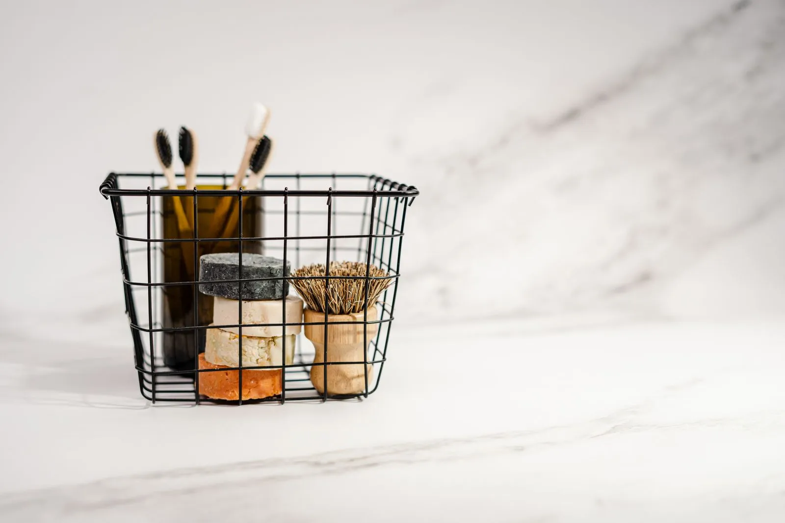 A small black wire basket holding two bamboo toothbrushes, unlabeled soap bars, and a small glass jar, sitting on a marble bathroom counter