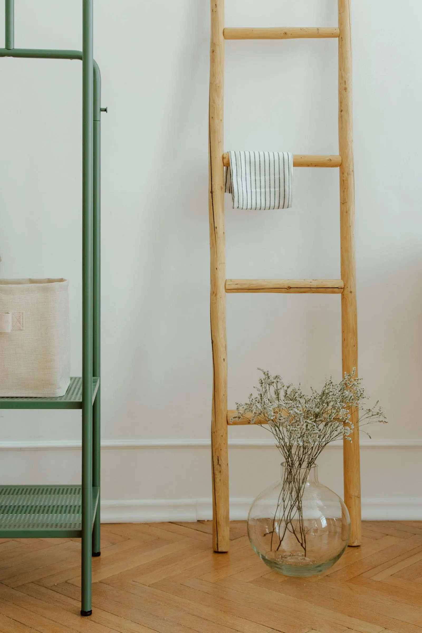 Wooden ladder-style shelf leaning against a wall with a striped cloth draped over one rung and a glass vase of dried flowers at its base