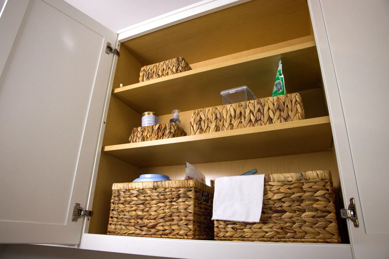 Open cabinet shelves holding woven storage baskets, a spray bottle, a jar, and a folded cloth