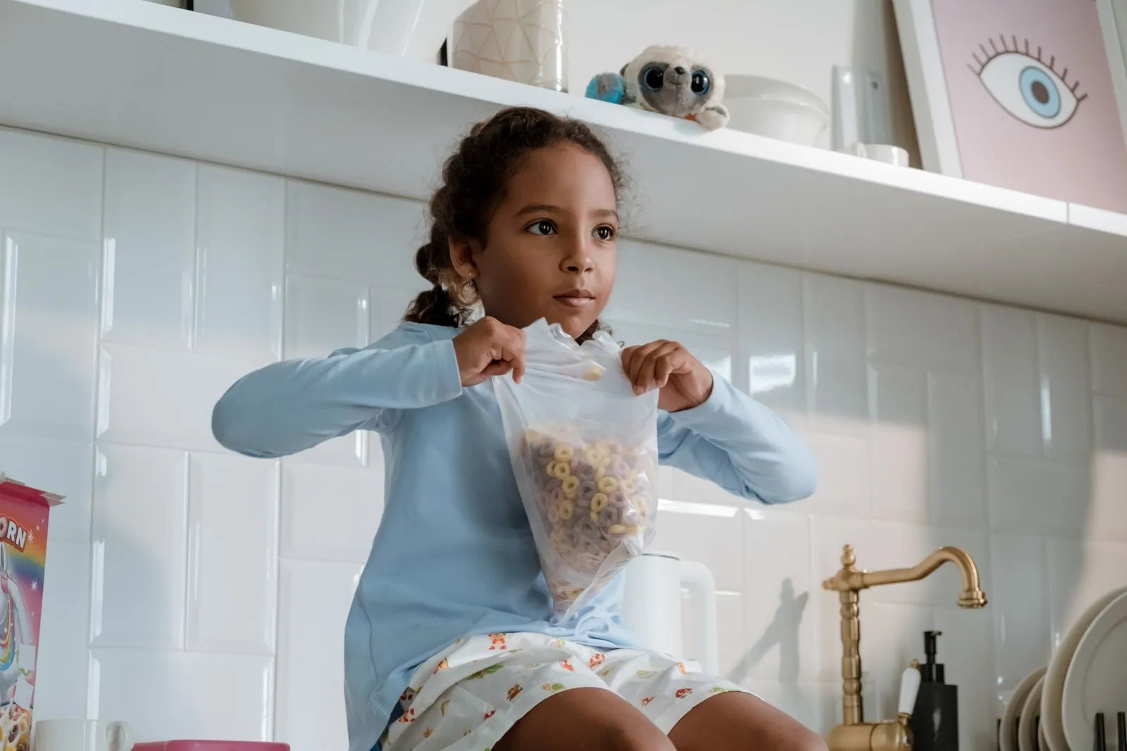 A child sitting in a kitchen holding an open bag of cereal, with a colorful unicorn-themed cereal box on the counter beside her