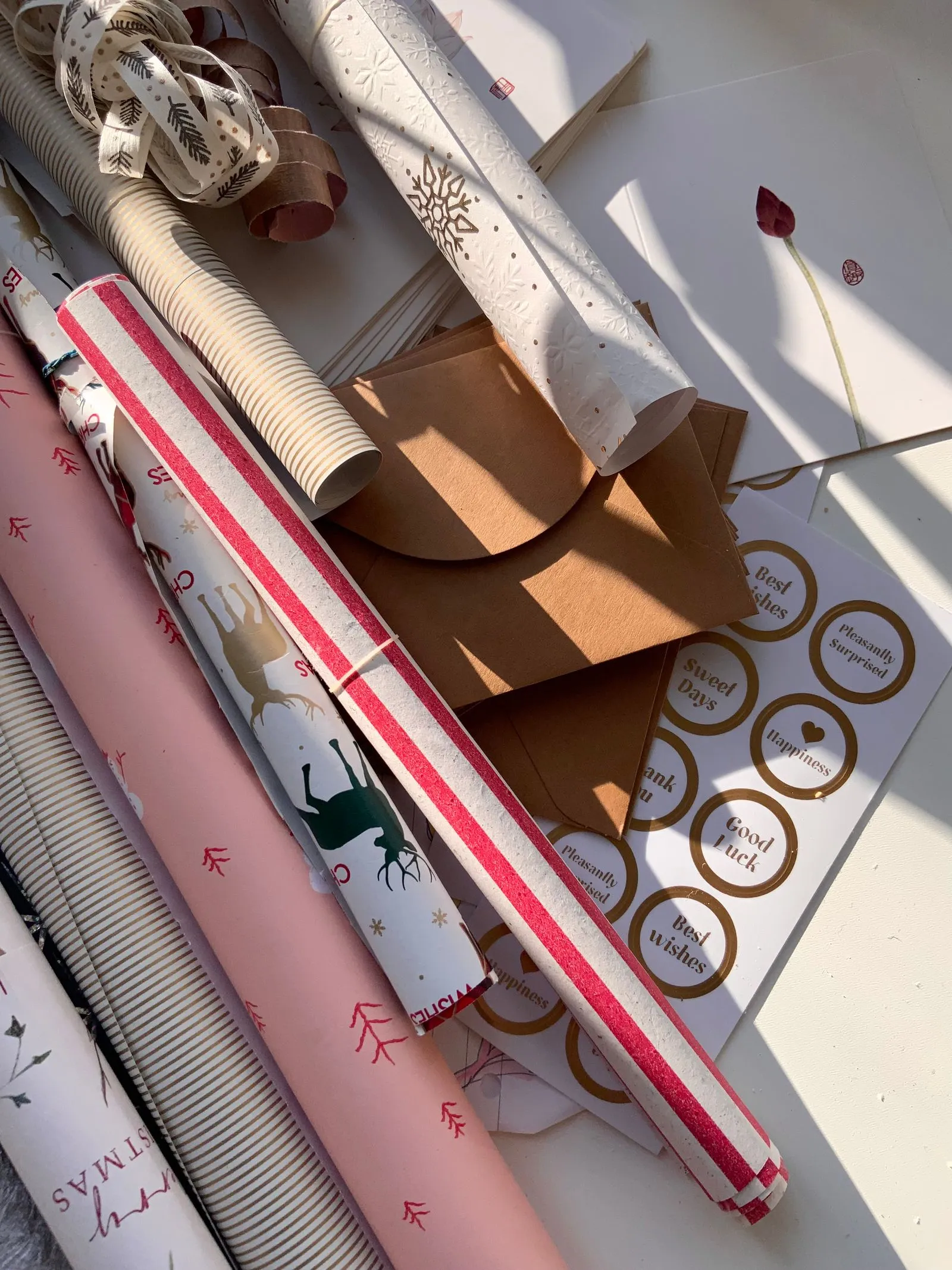 Overhead view of wrapping paper rolls, ribbon, kraft envelopes, and a sheet of gold circular gift tag stickers laid out in sunlight