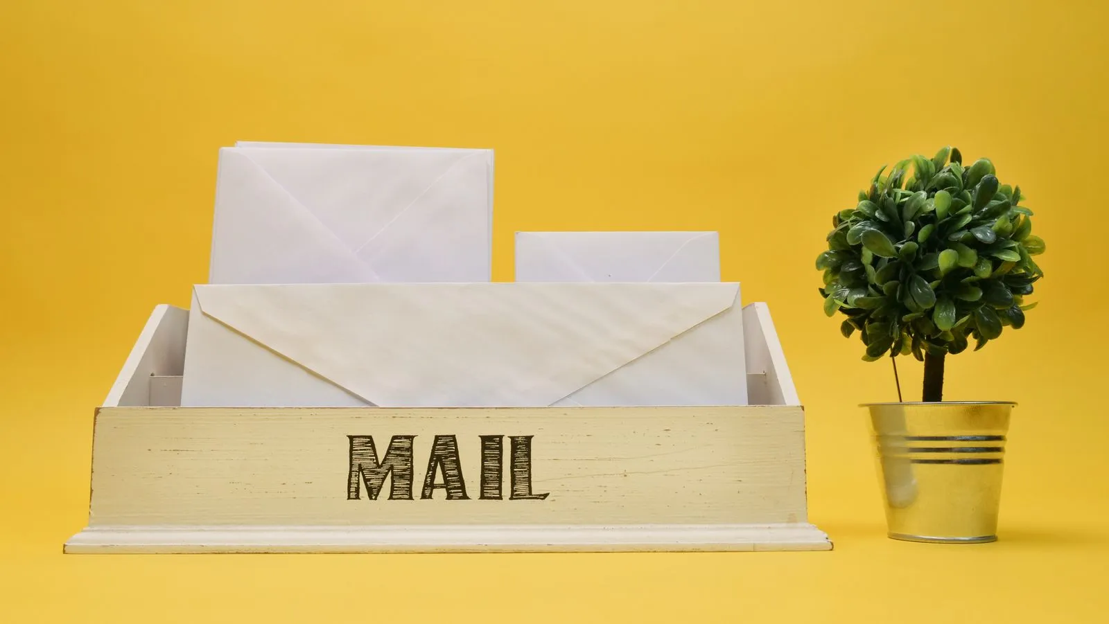 Wooden mail organizer tray holding white envelopes next to a small potted plant on a yellow background