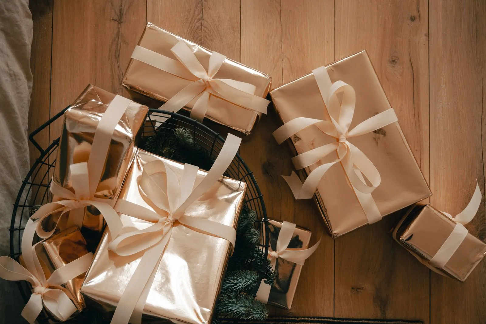 Overhead view of rose-gold wrapped gifts with cream ribbon bows piled inside a black wire basket alongside pine sprigs, on a wood floor