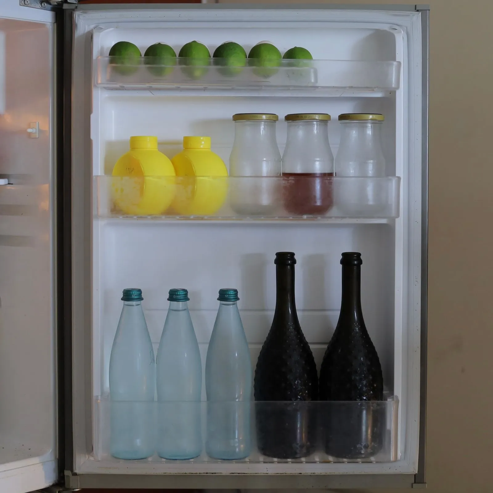 Open refrigerator door shelves neatly organized with limes on top, jars and bottles with yellow caps in the middle, and glass bottles below