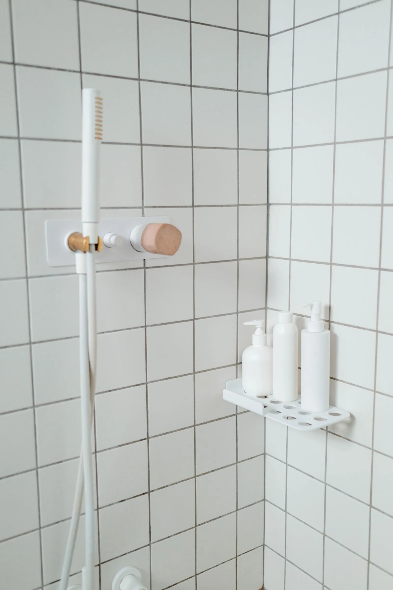 A white corner shower caddy mounted on tile holds three matching white pump bottles beside a wall-mounted shower control