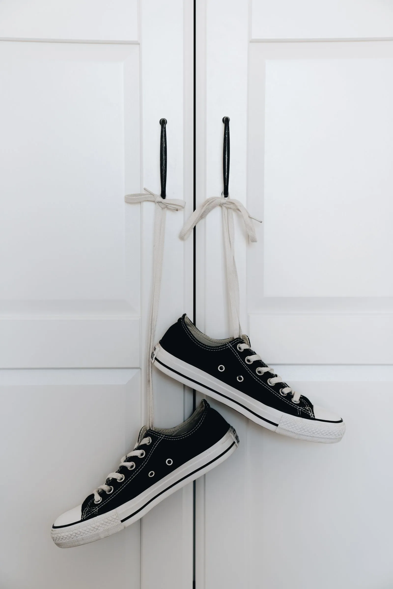 A pair of black canvas sneakers hangs by their own laces looped over two small hooks mounted on a white paneled door