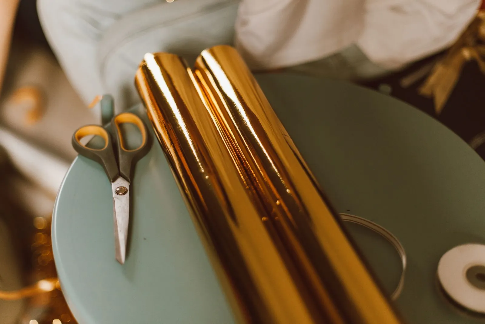 Rolled gold foil wrapping paper resting beside a pair of scissors and a ribbon spool on a small round side table
