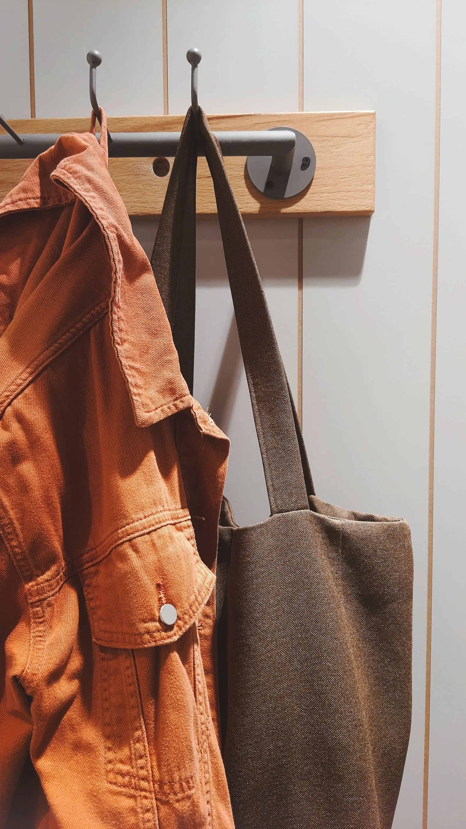 An orange denim jacket and an olive canvas tote bag hanging side by side from two wall-mounted hooks
