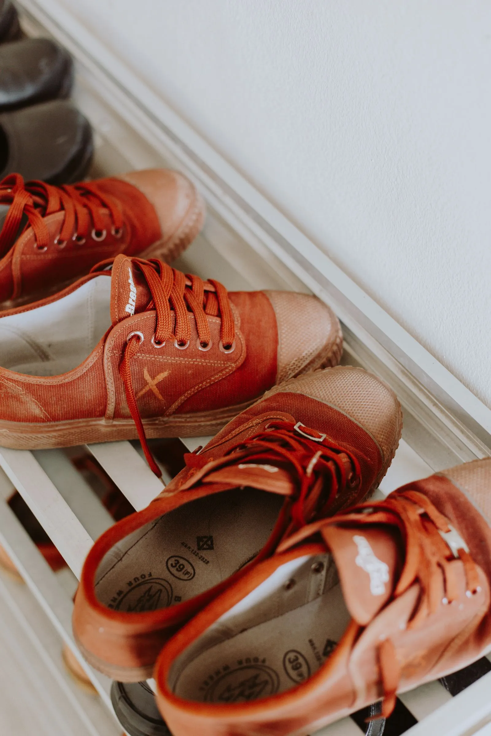 A close-up of orange canvas sneakers and slip-on shoes arranged in pairs on a white slatted shoe rack